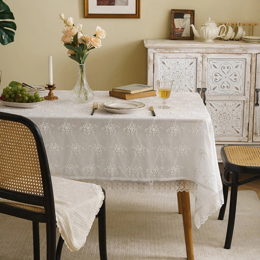 A white cotton tablecloth with European embroidery is laid out on a dining table, with intricate thread detailing visible. The table is set with cutlery and a book is placed on it, suggesting a sophisticated and elegant dining atmosphere.