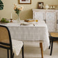 A white cotton tablecloth with European embroidery is laid out on a dining table, with intricate thread detailing visible. The table is set with cutlery and a book is placed on it, suggesting a sophisticated and elegant dining atmosphere.