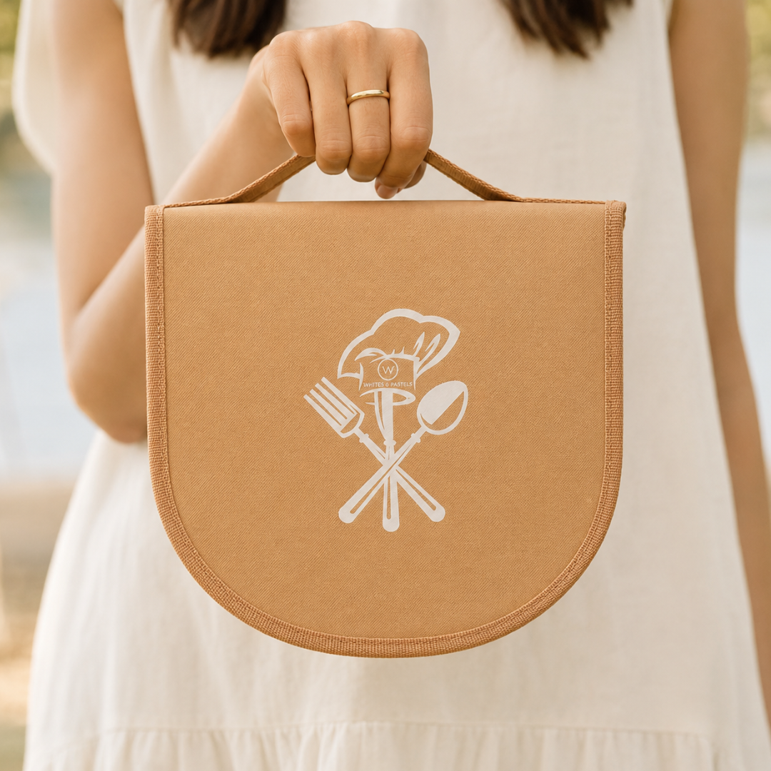 A person in a white sleeveless dress holds a tan fabric bag with a rounded bottom and top handle. The front has a white printed graphic of a chef's hat above crossed cutlery.