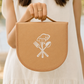 A person in a white sleeveless dress holds a tan fabric bag with a rounded bottom and top handle. The front has a white printed graphic of a chef's hat above crossed cutlery.