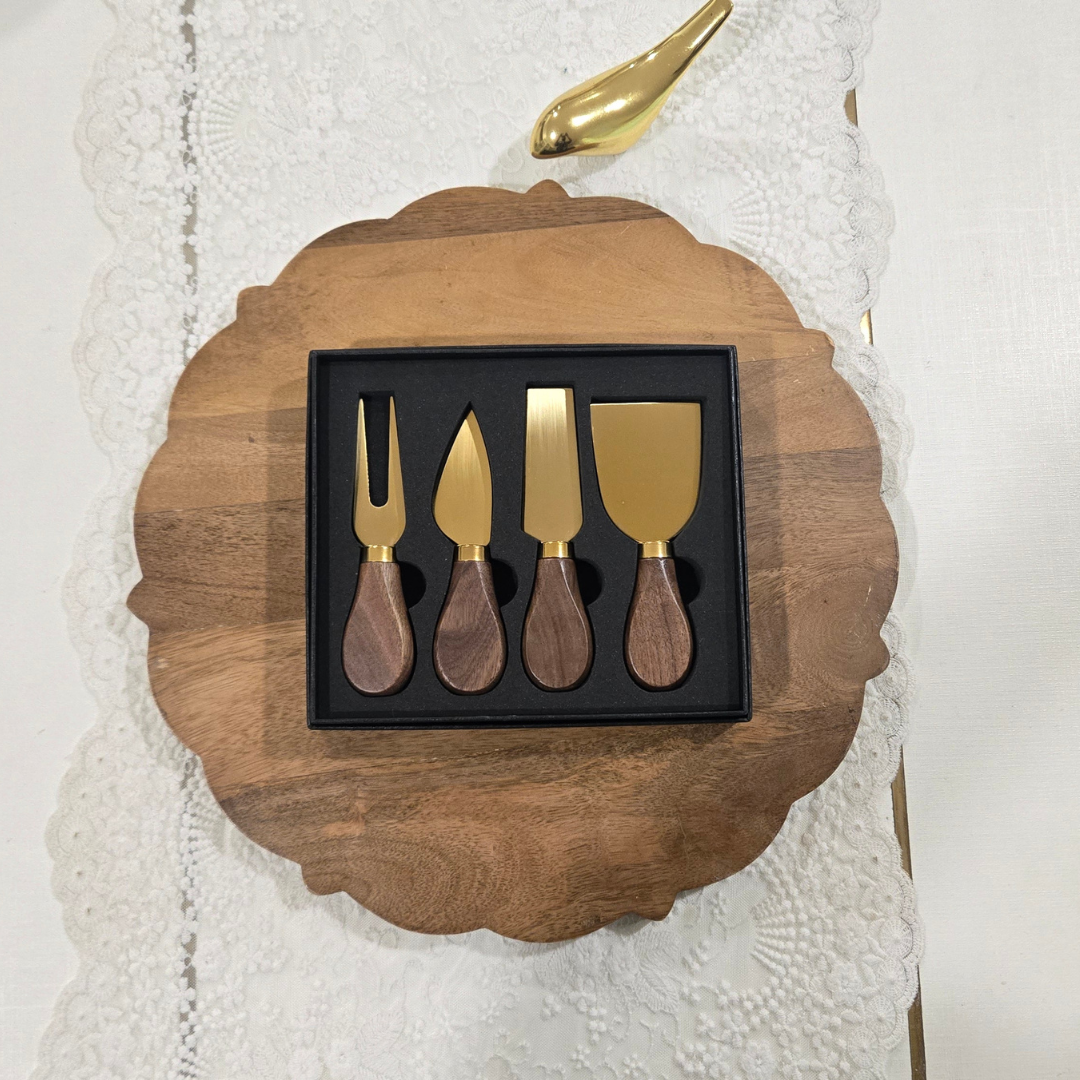 Four-piece cheese and carving utensil set with gold-tone metal heads and dark wood handles, presented in a black gift box on a scalloped wooden board over a white lace cloth.