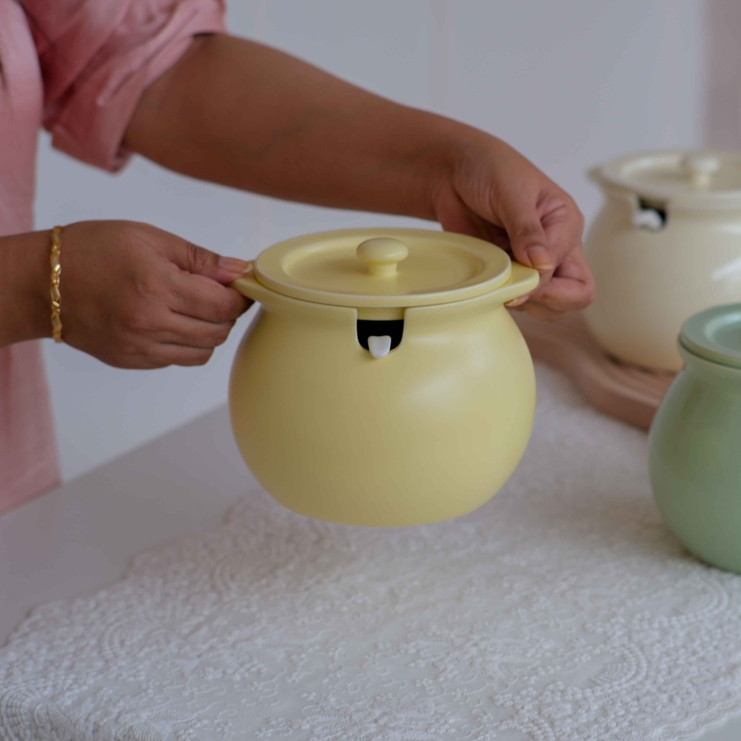 A person in a pink top holds a round matte yellow ceramic pot with a lid and small black handle detail, set on a white tablecloth with other pots in the background.