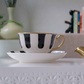 A white ceramic teacup and saucer set with bold black vertical stripes and gold trim, displayed on a white textured surface in a softly styled interior setting.