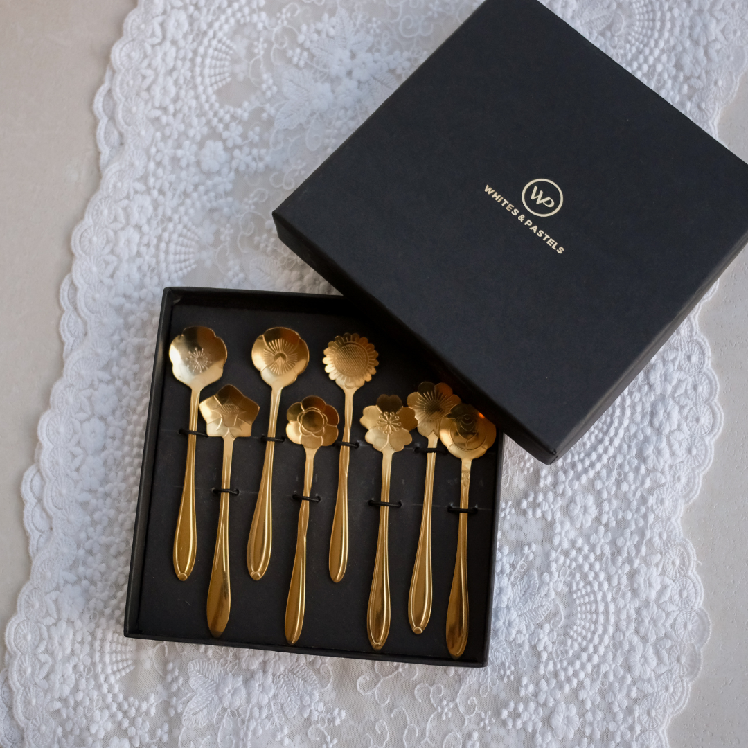 A set of eight gold-colored spoons with decorative floral and leaf-shaped bowls, presented in a black tray next to its black gift box on a white lace doily.