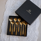A set of eight gold-colored spoons with decorative floral and leaf-shaped bowls, presented in a black tray next to its black gift box on a white lace doily.