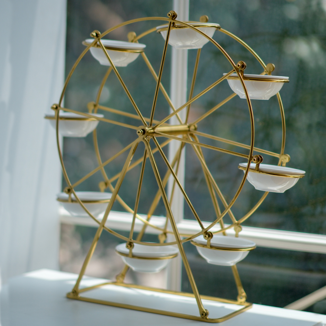 A gold-finished Ferris wheel tabletop decor piece with six white ceramic bowls, photographed on a windowsill with a soft-focus window background.