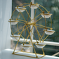 A gold-finished Ferris wheel tabletop decor piece with six white ceramic bowls, photographed on a windowsill with a soft-focus window background.