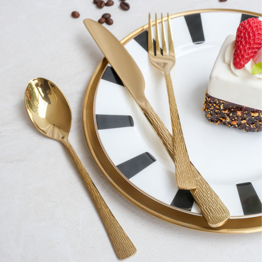 Gold-colored metal cutlery set including a spoon, knife, and fork, styled on a white plate with black stripes and gold trim alongside a slice of cake on a light textured surface.