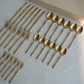 A set of matte gold flatware, including knives, forks, and spoons, arranged neatly on a light gray textured surface next to a beige box.