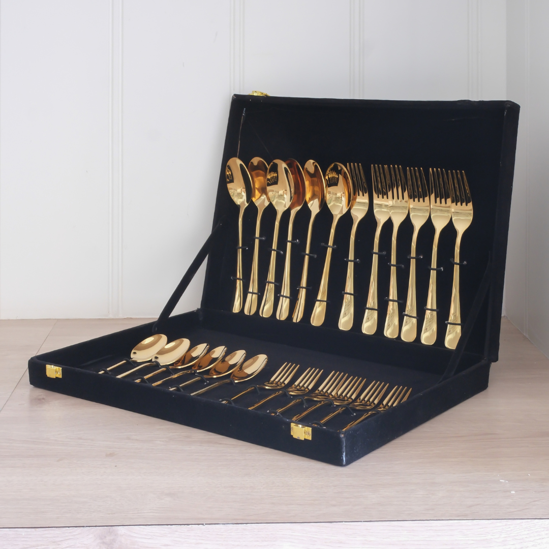 A gold flatware set with matching spoons and forks displayed inside an open black presentation case on a light wood surface against a white paneled wall.