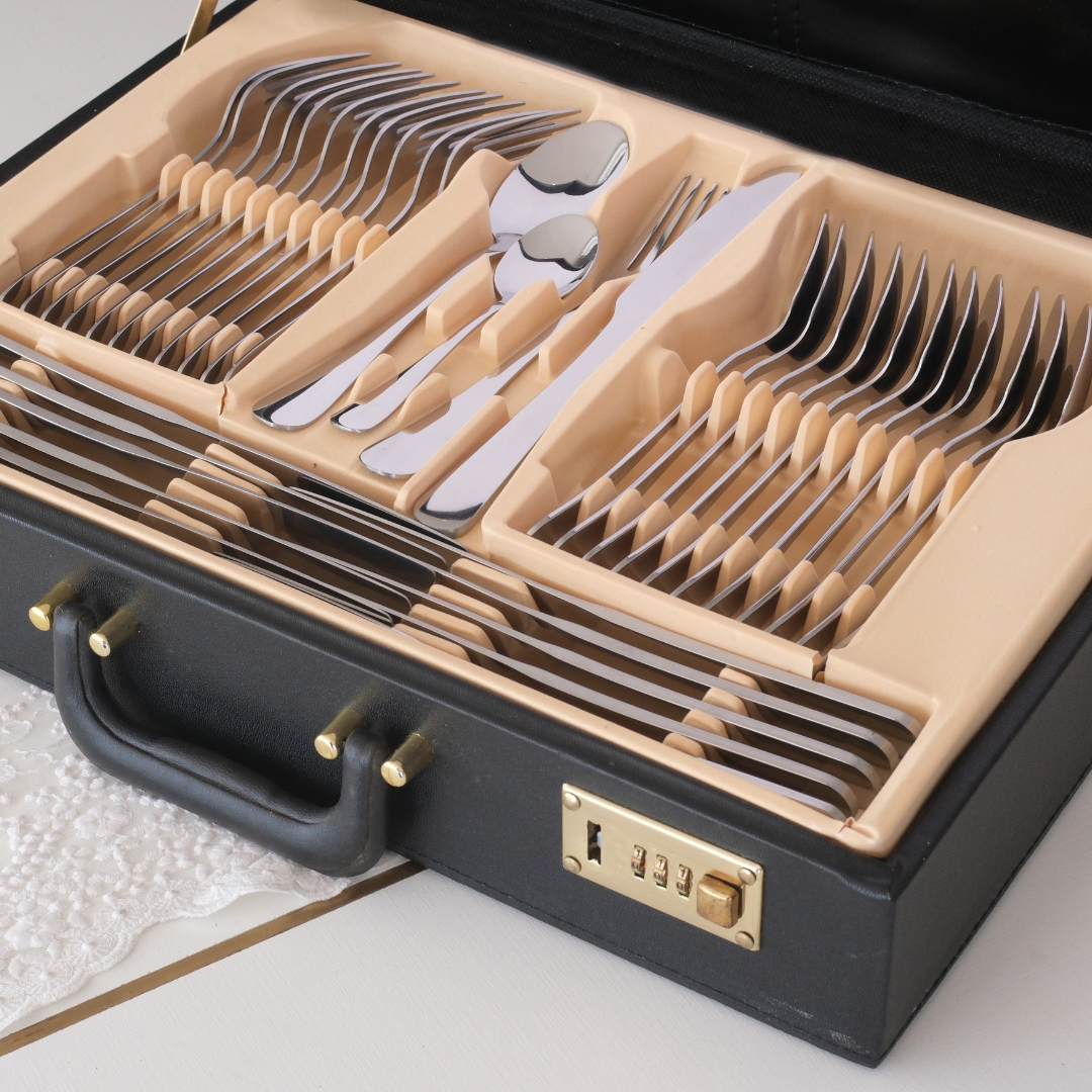 A black faux-leather briefcase-style storage box with complete set of silver cutlery consisting of Spoons, Forks and Dinner Knives resting on a white table runner in a home setting