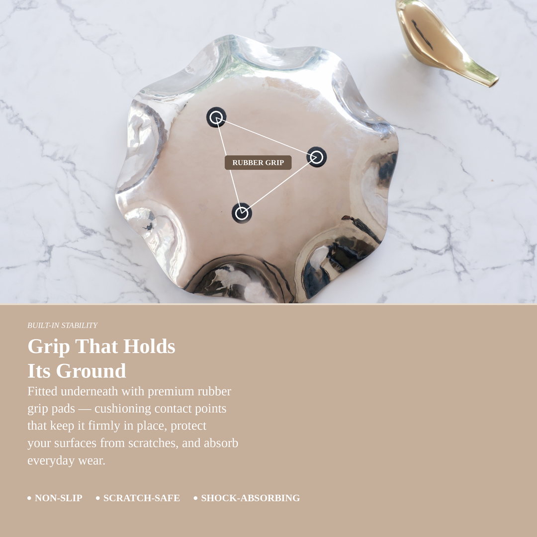 A metallic silver, scalloped-edge dish or coaster with built-in rubber grip pads underneath, shown on a white marble surface with a beige text banner below highlighting its non-slip, scratch-safe, and shock-absorbing features.