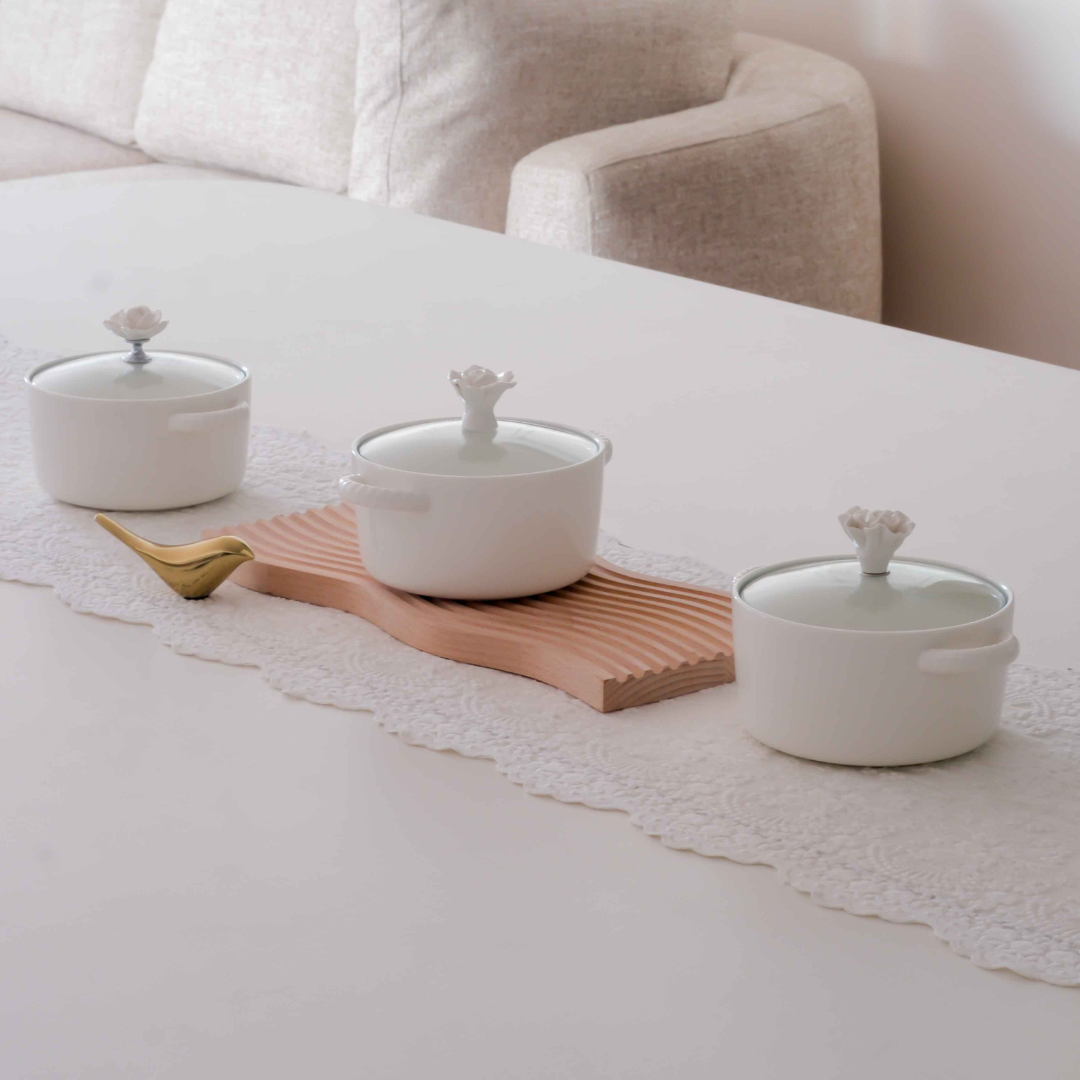 Three white ceramic pots with glass lid and decorative flowe shaped knobs are arranged on a white table runner with a wooden board and a small gold spoon, with a beige sofa in the background.