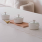 Three white ceramic pots with glass lid and decorative flowe shaped knobs are arranged on a white table runner with a wooden board and a small gold spoon, with a beige sofa in the background.